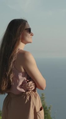Dikey video. Daire çizen sinematik kamera, dağın tepesinde genç bir kadın yakalıyor, günbatımının ve denizin manzarasının tadını çıkarıyor. Yavaş Çekimde.