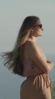 Dikey video. Ellerini bağlayan genç bir kadın, denizin arka planına karşı dağlarda gün batımını izliyor. Doğadaki yalnızlık ve sükunet kavramı..