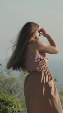 Dikey video. Genç bir kadının, arka planda mavi gökyüzü ile birleşen dağ zirvesinde dururken gün batımını seyrederken çekilmiş sinematik bir görüntüsü. Rüzgar yavaşça saçlarını okşuyor..
