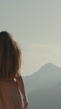 Dikey video. Genç bir kadın yüksek dağlarda yürür, sınıra yaklaşır ve dağın arkasında güneşin batışını takdir eder. Uzaktaki görkemli dağlar.