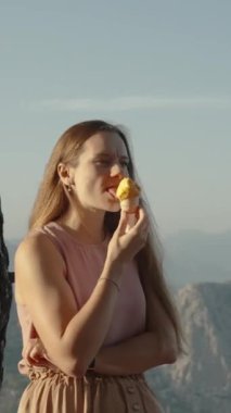Dikey video. Dağlardaki Güneş 'in batışından gelen sinematik ve destansı ışık, dondurma yiyen genç bir kadını aydınlatıyor ve dağların tepesinde eğlendiriyor..
