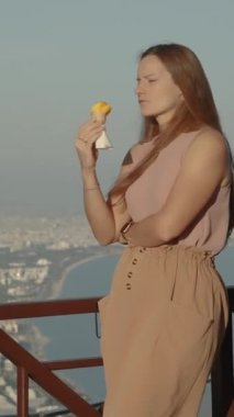 Dikey video. Dağın Tepesinde Genç Bir Kadının Sinematik Çekimi. Günbatımında Yumuşak Işık, Şehir ve Arka planda Deniz ile Dondurma Yiyor. Mountain Peak 'te bir kafe..