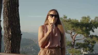 Uzun saçlı ve Avrupalı görünümlü genç bir kadın, günbatımında ağaçların tepesinde dikilirken dondurma yiyor..