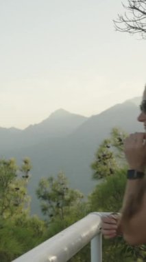 Dikey video. Gezgin genç kadın uçurumun kenarına yaklaşıyor. Tırabzanlara yaslanarak, dağların ve denizin güzel manzarasına hayran. Gün batımında sinematik ışık.