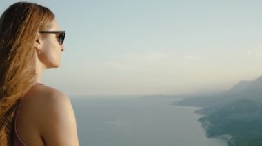 Dağın en yüksek noktasında duran genç bir dişi turist denizin üzerinde gün batımını izliyor. Panoramik görünümde sinematik ve destansı ışıklandırma.