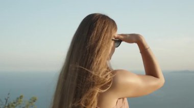 Gün batımından gözleri kamaşmış genç bir kadın dağın zirvesinde duruyor ve denize ve kayalıklara bakarken elleriyle gözlerini koruyor..