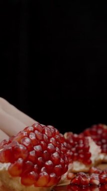 Dikey video. Masanın üzerinde olgun ve sulu narlar sergileniyor ve arka planda bir parça yiyen genç bir kadın var. Yavaş çekim.