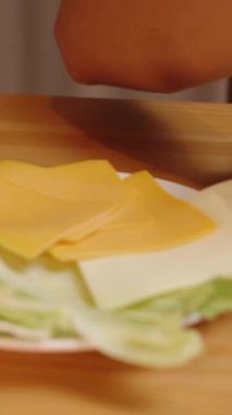 Dikey video. Peynir Dilimleri Bir Kadın Tarafından Burger Buns 'a yerleştirilir. Yakınlarda düzgünce hazırlanmış sebzeler ve diğer malzemeler var. Sıcak bir ev atmosferi ve güzel bir iç mekan..