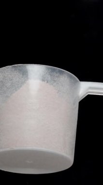 Dikey video. Siyah bir arka planda, ölçüm kaşığından etrafa protein tozu serperken en üst görüntü. Spor beslenme konsepti. Yavaş çekim makrosu.