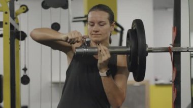 Spor salonundaki genç kadın, terli halterler içinde halter için ağırlık tasması takmaya çalışıyor. Çok sıkı ve zar zor idare ediyor..