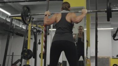 Kalça kasları gelişmiş genç bir kadın spor salonundaki aynanın önünde halterle çömeliyor. Arka plan, yavaş çekimde.