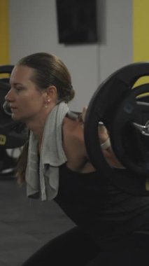 Dikey video. Genç atletik kadın omuzlarında ağır bir halterle çömelme hareketleri yapıyor, ayaklarının altında destek için küçük ağırlık plakaları kullanıyor..