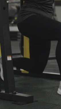 Dikey video. Sırılsıklam olmuş genç bir kadın, tek bacaklı bir makine üzerinde çömelme hareketi yapıyor. Bir Yumuşak Yastık Boyun Desteği için Halterin üzerine yerleştirilmiştir.
