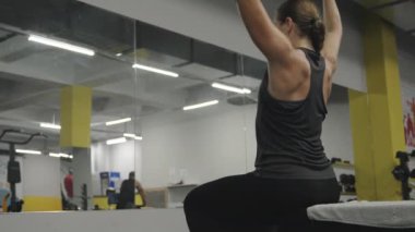 Genç bir kadın spor salonundaki bir bankta oturuyor, dambıl kaldırıyor, aynanın önünde, doğru infazı izliyor..