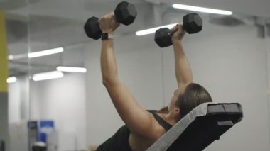 Spor salonunda göğüs kasları için egzersiz yapan bir kadın. Dambıllarla kollarını yana açıyor..