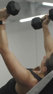 Dikey video. Spor salonunda göğüs kasları için egzersiz yapan bir kadın. Dambıllarla kollarını yana açıyor..