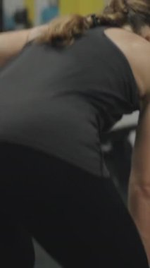 Dikey video. Güçlü ve bağımsız genç bir kadın tek eliyle ağır bir dambılı kaldırıyor. Spor salonunda dayanıklılık eğitimi..