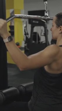 Dikey video. Genç bir kadın geniş kabzalı geniş omuzlu latissimus dorsi kaslarını ve omuz kemerini geliştiriyor..