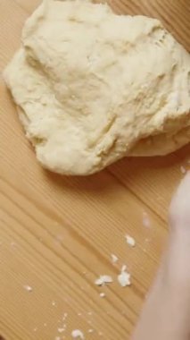 Dikey video. Tahta bir ev masasının arkasında hamuru elle yoğuran bir kadın. Mutfak aletleri masanın üzerine saçılmış. Kamera ağır çekimde döner..