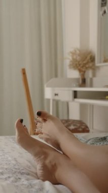 Dikey video. Güneşli bir sabahta parlak yatak odası. Kara Tırnak Cilalı Kadın Ayakları Yatakta Yatarken Parmaklarını Kıpırdatıyor. Rüzgâr Perdeleri Hışırdatır.