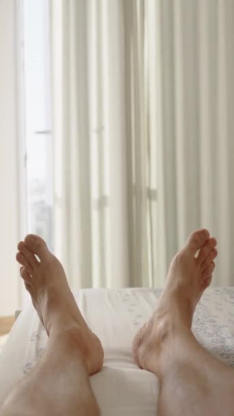 垂直录像 一个男人躺在一个阳光明媚的卧室里 把一条腿放在另一条腿上 第一人称视图 — 图库视频影像