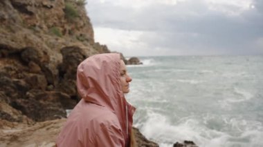 Pembe paltolu genç bir kadın uzaklığa bakıyor, düşüncelere dalmış. Yüksek dalgalar kayalara çarpar ve o da kenarlarda oturur. Yavaş Hareket.