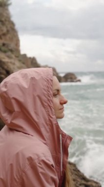 Dikey video. Pembe paltolu genç bir kadın uzaklığa bakıyor, düşüncelere dalmış. Yüksek dalgalar kayalara çarpar ve o da kenarlarda oturur. Yavaş Hareket.