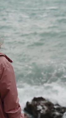 Dikey video. Pembe yağmurluk giyen yalnız bir genç kadın bir deniz fırtınasında kayalıklarda dikiliyor ve uzaklığa bakıyor. Yavaş Çekimde.