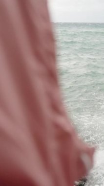 Dikey video. Pembe kapüşonlu bir yağmurluğun arkasından üçüncü şahıs manzarası, denizdeki fırtınayı ve yüksek dalgaları gözlemler. Yavaş Çekimde.