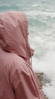 Dikey video. Pembe yağmurluklu genç bir kadın, Deniz Fırtınası nedeniyle dalgaları gözlemler. Suyun kenarındaki kayalıklarda duruyor ve damlacıklar ona ulaşıyor. Yavaş Çekimde.