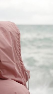 Dikey video. Kamera kapüşonlu genç bir kadından yavaşça uzaklaşır, pembe bir yağmurluk giyer, kayaların üzerinde durur ve deniz fırtınasını gözlemler..