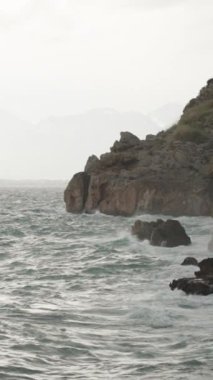 Dikey video. Kaynar Deniz ve Bulutlu Hava, Uzak Dağlar ve Yüksek Kayalıklara Su Sıçrayan Su. Yavaş Hareket.