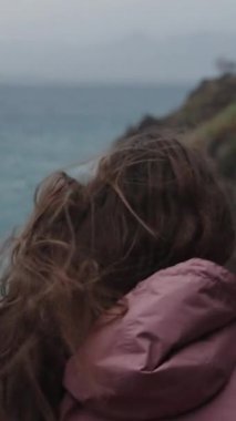 Dikey video. Yükseklerdeki Genç Bir Kadın, Denizdeki Fırtına, ve Güçlü Rüzgar Uzun Saçlarını ve Pembe Yağmurluğunu Kaldırıyor, Arkadan Görünüşü.