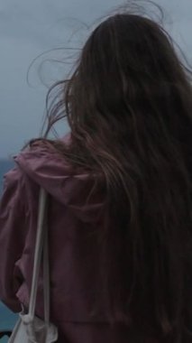 Dikey video. Deniz kıyısındaki bir uçurumun kenarında duran yalnız kadın, Fırtına ve Güçlü Rüzgar, Uzun Saçlarını Uzağa Bakarken.