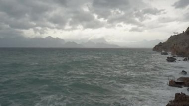 Denizde bulutlu hava, güçlü rüzgarlar arka planda büyük dalgalar, kayalar ve dağlar oluşturur..