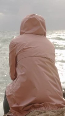 Dikey video. Pembe yağmurluklu bir kadın uçurumun kenarında oturur, bulutlu hava ve rüzgar, ama güneş ışınları bulutları aşar, ve dalgalardan gelen su damlaları neredeyse ona ulaşır.