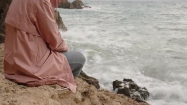 Pembe ceketli, başlıklı yalnız bir kadın kayanın üzerinde oturur ve öfkeli denize bakar..