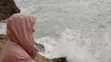 Güçlü Rüzgarda Deniz kenarındaki Kayaların Arasında Pembe Kapüşonlu Yağmurluk Giyen ve Parmağıyla Bir Yeri Gösteren Bir Kadın.