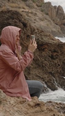 Dikey video. Yağmurluk giyen bir kadın, telefonunda Fırtınalı Deniz 'in Fotoğrafını Çekiyor..