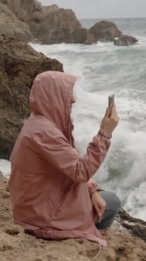 Dikey video. Pembe Kapüşonlu Genç Bir Kadın Denizde Büyük Dalgaların Fotoğraflarını Çekiyor, Keskin Kayalarda Otururken, Püskürtücüler Neredeyse Ona Ulaşırken.