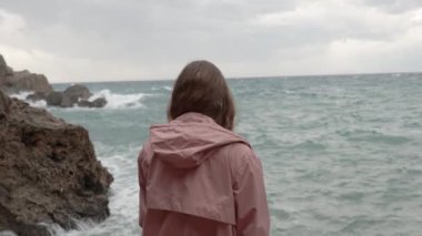 Yalnız bir kadın Öfkeli Deniz 'e bakıyor ve Dalgalar Keskin Kayalara Çarpıyor. Yavaş Hareket.