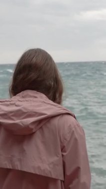 Dikey video. Yalnız bir kadın Öfkeli Deniz 'e bakıyor ve Dalgalar Keskin Kayalara Çarpıyor. Yavaş Hareket.