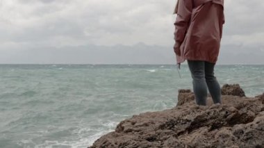 Pembe Yağmurluk Giyen Bir Kadın Deniz kenarındaki bir uçurumun kenarında duruyor ve Fırtınayı izliyor. Yavaş Hareket.