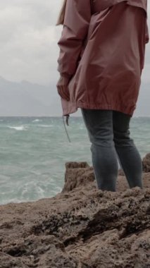 Dikey video. Pembe Yağmurluk Giyen Bir Kadın Deniz kenarındaki bir uçurumun kenarında duruyor ve Fırtınayı izliyor. Yavaş Hareket.