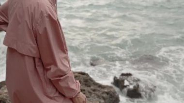 Kamera keskin kayaları ve suyun kenarında duran genç bir kadını yakalıyor. Denizde güçlü dalgaları olan bir fırtına var. Yağmurluk giyiyor..