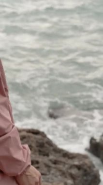 Dikey video. Kamera keskin kayaları ve suyun kenarında duran genç bir kadını yakalıyor. Denizde güçlü dalgaları olan bir fırtına var. Yağmurluk giyiyor..