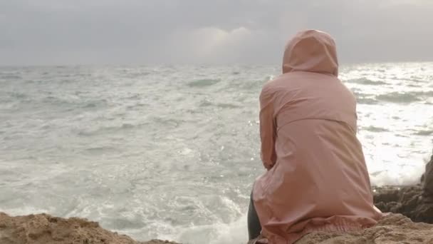 ピンクのレインコートの女性は クリフの端に座って 天気と風をオーバーキャストしますが 太陽は雲を介して壊れ 波からほぼ彼女にスプラッシュします — ストック動画