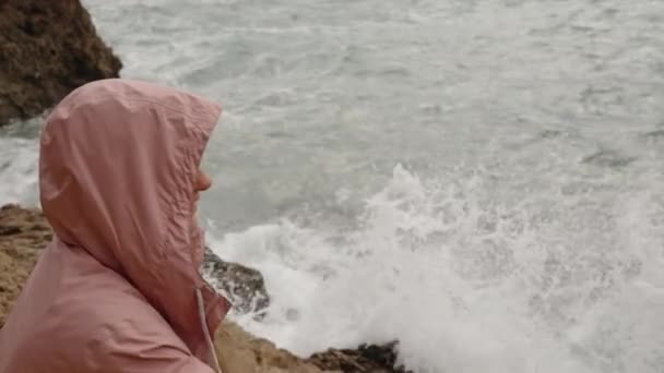 一个女人站在海边的岩石中 身披粉色雨衣 带着手指指向某个地方 — 图库视频影像