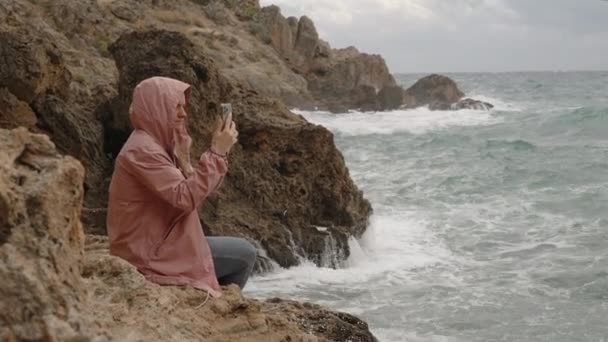 一个穿着雨衣的女人坐在锐利的岩石上 在她的手机上拍了一张风暴海的照片 — 图库视频影像