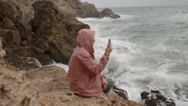 一位穿着粉红雨衣的年轻女子坐在锋利的岩石上 坐在海浪中 喷出的浪花几乎到达了她的身边 — 图库视频影像